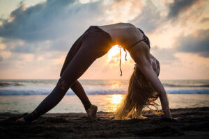 A woman in wheel pose with setting sun and ocean waves behind her